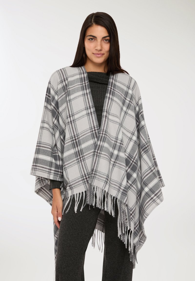 Donna che indossa un poncho a frange grigio a quadretti sopra un maglione scuro a coste e pantaloni, in piedi contro uno sfondo bianco semplice.