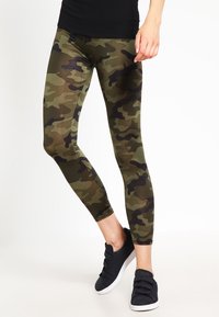 Camo leggings featuring green and brown patterns, made from stretchy material, with a fitted design and a smooth texture. Paired with black sneakers.