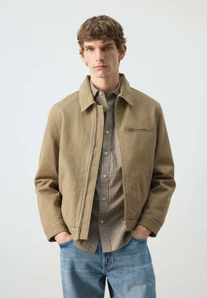 Hombre joven con chaqueta beige con cremallera sobre camisa a cuadros y vaqueros azules, de pie con las manos en los bolsillos contra un fondo liso.