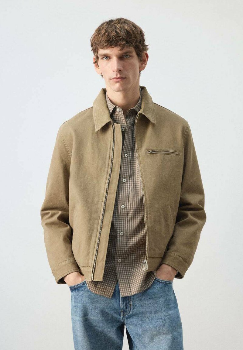 Mladi moški, ki nosi bež jakno z zadrgo čez karo srajco in modre kavbojke, stoji z rokami v žepih na enobarvnem ozadju.