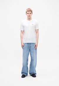 Hvit t-skjorte med korte ermer og liten svart logo, kombinert med lys blå baggy denimjeans og svarte sko. Ensfarget hvit bakgrunn.