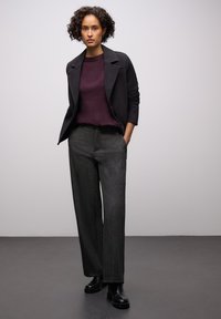 Zwarte op maat gemaakte jas over een bordeauxrode trui, gecombineerd met grijze wijde broekspijpen en zwarte enkellaarsjes. Eenvoudige, strakke lijnen en gestructureerde stof.