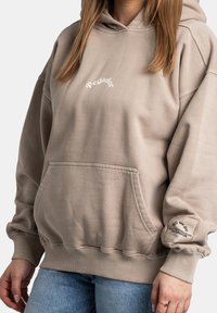 Sudadera beige con capucha fabricada en tejido suave, que presenta un bolsillo frontal, puños acanalados y un logotipo en el pecho y la manga. Ajuste casual.