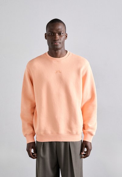 Sweatshirt de culoare piersică, guler rotund, material moale, manșete și tiv riglate, logo mic pe piept, purtat cu pantaloni gri.