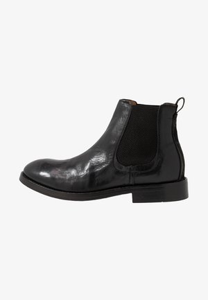 Bota Chelsea negra de cuero con paneles laterales elásticos, punta redonda y acabado texturizado. Presenta una suela plana y un tirador en el talón.