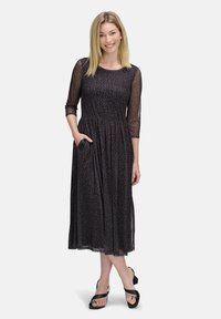 Maxikleid aus schwarzem Stoff mit floralem Muster, halblangen transparenten Ärmeln, gerafftem Bund und seitlichen Taschen. Trägt dazu schwarze Sandalen.