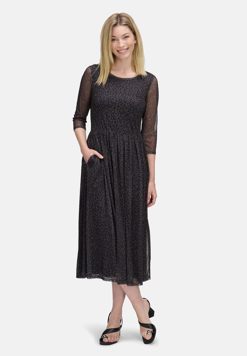 Maxikleid aus schwarzem Stoff mit floralem Muster, halblangen transparenten Ärmeln, gerafftem Bund und seitlichen Taschen. Trägt dazu schwarze Sandalen.