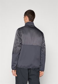 Temno siva jakna z gladkim, sijočim zgornjim delom in teksturiranim spodnjim delom. Ima stoječ ovratnik in elastične manšete.