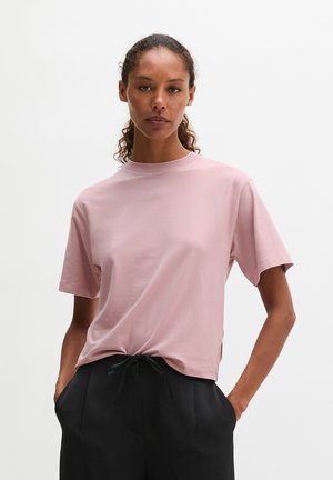Vrouw met krullend haar die een lichtroze T-shirt met korte mouwen draagt, lichtjes ingestopt in zwarte broek met koordjes, staat met handen in haar zakken.