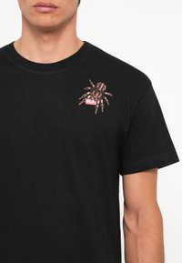 T-shirt en coton noir avec un graphisme d'araignée et le mot "DIESEL" en rouge, situé sur le côté gauche de la poitrine. Manches courtes et col rond.