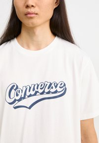 Hvit bomull t-skjorte med stor blå grafisk tekst "Converse" over brystet. Korte ermer, rund hals. Jevn tekstur.