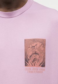 Κοντινό πλάνο ενός ροζ πουκάμισου με ένα σκούρο σχέδιο που απεικονίζει ένα χέρι και ένα ζώο περιβαλλόμενα από φυτά και κείμενο που λέει "ΣΥΓΚΡΟΥΣΗ. ΑΝΘΕΞΗ. ΑΝΑΔΥΣΗ. ΜΙΑ ΕΡΓΑΣΙΑ ΣΕ ΕΞΕΛΙΞΗ."