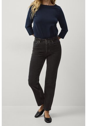 Woman wearing a navy long-sleeve top, high-waisted black jeans, and black flats, standing with hands behind her back.