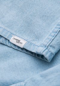 Tissu en denim bleu clair avec une texture douce ; comporte une étiquette cousue portant la mention "OMBRA WEAR" en noir sur fond blanc.