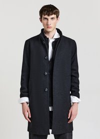 Cappotto lungo in lana nera con colletto, cinque bottoni frontali e tasche laterali. Il modello indossa una camicia bianca sotto, con le maniche arrotolate.