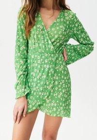 Femme portant une robe longue à manches longues à fleurs vertes avec des poignets volantés, posant avec une main sur la hanche et l'autre reposant sur sa cuisse.