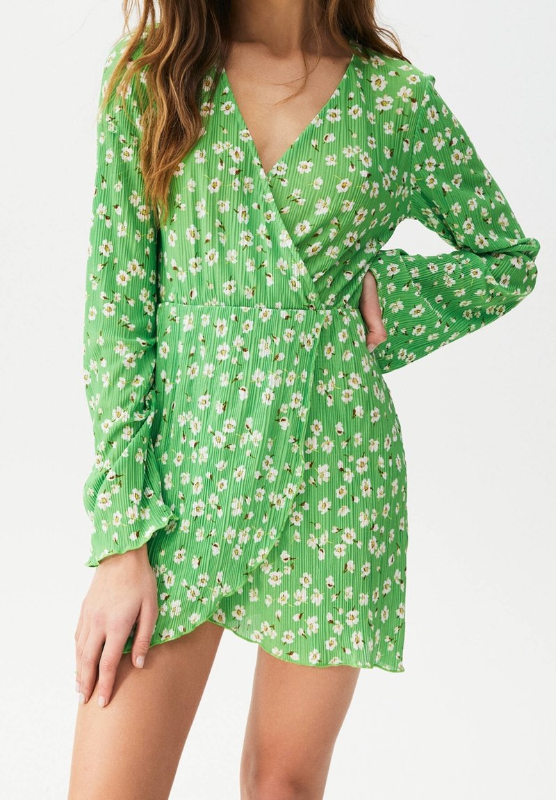 Femme portant une robe longue à manches longues à fleurs vertes avec des poignets volantés, posant avec une main sur la hanche et l'autre reposant sur sa cuisse.