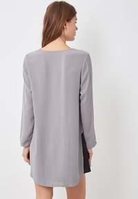 Mujer con el cabello largo y castaño de espaldas, vistiendo un vestido gris suelto de manga larga con un panel lateral negro sobre un fondo blanco.