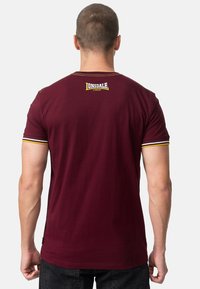 Bordo medvilnės marškinėliai su geltonomis ir baltomis juostelėmis ant rankovių, su logotipo etikete nugaros kaklo zonoje. Klasikinis siluetas.