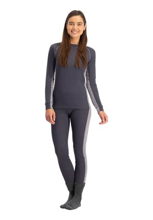 Lachende vrouw staand, gekleed in een donkergrijze top met lange mouwen en bijpassende leggings met lichtgrijze zijpanelen, en donkergrijze sokken.