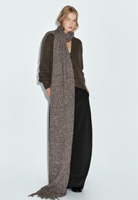 Lange, gebreide sjaal in een mix van bruin en grijs tinten, met een gestructureerd patroon en franjes aan de randen, gecombineerd met een donker oversized cardigan en broek.