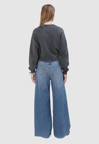 Person mit langen Haaren, die einen dunkelgrauen Pullover und weit geschnittene blaue Jeans trägt, steht mit dem Rücken zu sehen vor einem einfarbigen grauen Hintergrund.