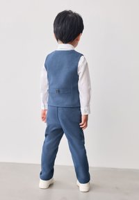 Vista trasera de un niño con un traje azul a medida de tela texturada, camisa blanca y deportivas claras, con un detalle de hebilla en la cintura.
