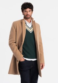 Beige kappor med en strukturerad krage, bärs över en mörkgrön tröja med V-ringning och krämfärgade ränder, ihop med mörka jeans.