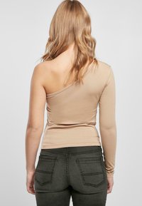 Beige en-sides topp i myk stoff, tettsittende design, lang erme på den ene siden, sammen med mørke grå jeans med baklommer.