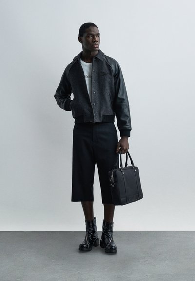 Άνδρας με μαύρο και γκρι bomber μπουφάν, λευκό πουκάμισο, μαύρα φαρδιά κοντά παντελόνια μέχρι το γόνατο, μαύρα μπότες με τακούνι, κρατώντας μαύρη τσάντα χειρός, στέκεται σε γκρι πάτωμα.