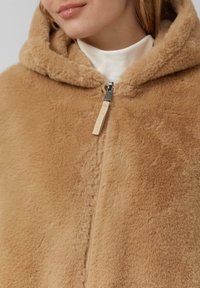 Tan nepbont capuchoncape met een gladde textuur, vooraan met een ritssluiting en een leren merklabel. Beschikt over zachte, warme stof met een knusse uitstraling.