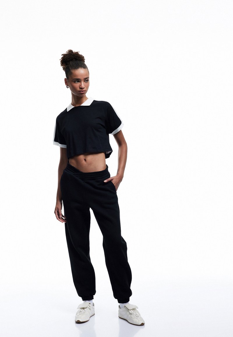 Sort cropped t-shirt med hvid krave og manchetter, parret med sorte joggers. Fodtøj er hvide sneakers med sløjfe-detaljer.