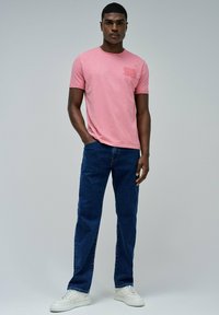Camiseta rosa de manga corta con un logo sutil en el pecho, combinada con jeans de mezclilla azul y zapatillas blancas. Atuendo sencillo y casual.