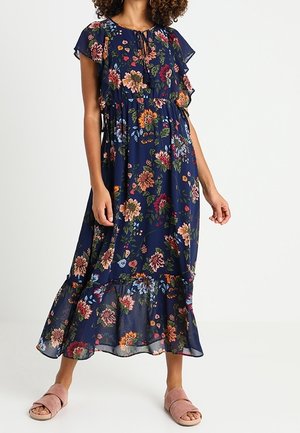 Nainen pukeutuneena tummansiniseen kukkakuosiseen midimekkoon, jossa on röyhelöhihat ja helma, yhdistettynä vaaleanpunaisiin avokkaisiin, seisoo valkoista taustaa vasten.