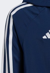 Veste de sport marine avec des bandes latérales blanches, texture lisse et logo brodé. Comprend une capuche et un design structuré.