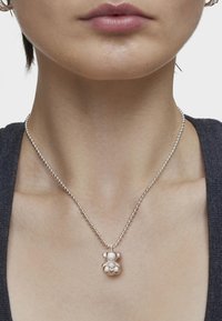Collar de bola de cadena de plata con un colgante de osito de peluche con acabado brillante y acentos de perlas, descansando sobre un escote neutro.