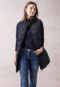 Svart lätt vadderad väst med hög krage, dragkedjestängning och axelsömmar, bärs över en marinblå tröja och blå jeans, stående mot en neutral bakgrund.