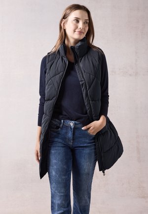 Zwarte gebreide vest met hoge kraag, ritssluiting en schouderzomen, gedragen over een marineblauwe trui en blauwe jeans, staande tegen een neutrale achtergrond.