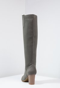Bottes grises montant jusqu'au genou avec un design perforé, une texture lisse et un talon empilé. Elles présentent un bout arrondi et une languette de traction en haut.