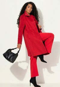 Rode wollen jas met een kraag, gecombineerd met rode broek en zwarte hoge hakken. Model houdt een zwarte leren handtas vast.