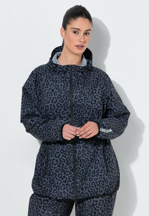 Vrouw draagt een donkergrijze leopardprint capuchonjas met een rits aan de voorkant en bijpassende broek, staand tegen een witte muur.