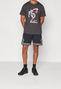 Man in zwarte basketbalschoenen en een donkergrijze t-shirt met een wit en rood basketbalontwerp, zwarte sokken en zwarte sneakers.