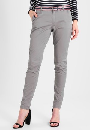 Femme portant un pantalon gris slim, des talons blocs ouverts noirs, un haut à manches longues rayé et une ceinture rayée rose et blanche.