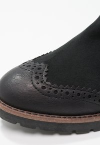 Bottine en cuir noir avec suède texturé, présentant un design perforé, des accents en zigzag, et une semelle épaisse en caoutchouc avec un bord marron contrastant.