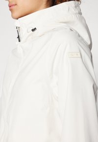 Veste imperméable blanche avec un col montant, une capuche ajustable et des accessoires noirs. Présente un patch logo crème sur l'épaule gauche.