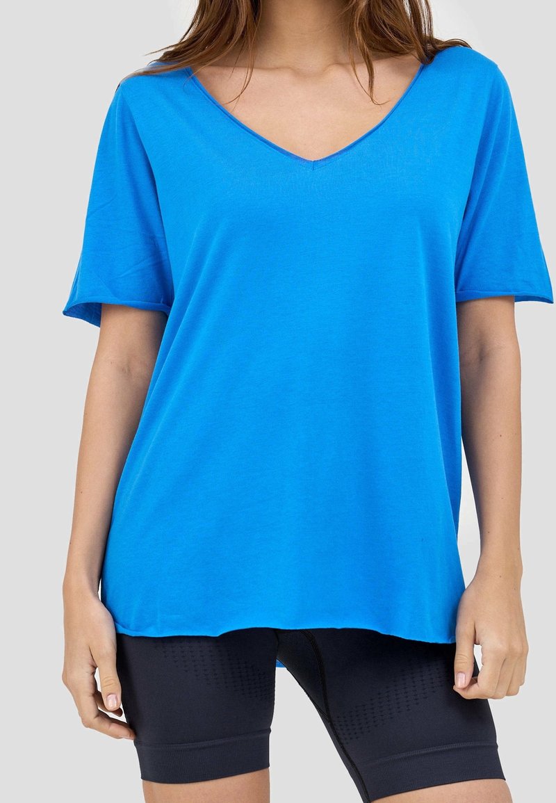 Femme portant un t-shirt bleu ample à col en V et manches courtes et un short de cycliste noir ajusté, debout devant un fond uni.