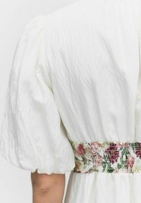 Weißes Kleid mit Puffärmeln, das über ein floral gemustertes Taillenband mit rosa und grünen Akzenten verfügt und aus einem strukturierten Stoff gefertigt ist.