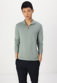 Light green quarter-zip sweater made of soft material, featuring long sleeves and ribbed cuffs and hem. Worn with dark pants.