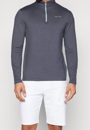 Mann trägt ein dunkelgraues Langarm-Top mit Viertelreißverschluss und Calvin Klein-Logo sowie weiße Knie kurze Shorts.