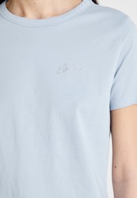 Light blue cotton t-shirt with a crew neckline and short sleeves, featuring a small, crystal-embellished logo on the left chest.